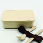 Closed beige wheat-straw lunch box displayed with matching fork, knife and textured brown elastic retaining strap.