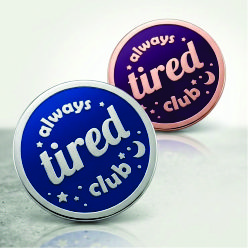 Two round 'Always Tired Club' enamel pins in blue and purple with silver lettering, displayed on a clean surface.