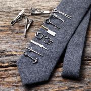 Collection of silver novelty tie clips shaped like scissors, keys, glasses, tools and an anchor displayed on a grey tie.